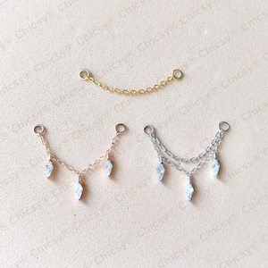 May include: Jewelry components in gold, rose gold, and silver tones. The image shows a gold chain with two loops, and two other chains with three dangling crystal-like charms. The background is a light pink color.