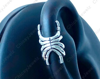 May include: A silver ear cuff with a skeletal design. The cuff wraps around the ear, featuring a series of curved, rib-like structures. The jewellery is displayed on a black ear-shaped form, highlighting its intricate details.