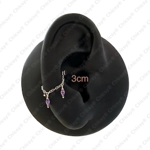 May include: A silver chain earring with two dangling purple teardrop-shaped gems. The earring is attached to a black ear model. The text "3cm" is visible on the image, indicating the length of the earring.