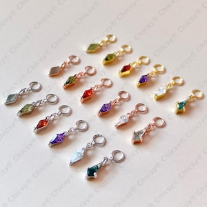 May include: A selection of small charms with diamond-shaped gemstone pendants in various colours. The metal findings are gold, silver, and rose gold. The charms include red, purple, green, and clear gemstones. The charms are displayed on a light-coloured surface.
