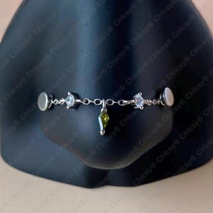 May include: A silver-toned nose cuff featuring a delicate chain with clear and green gemstone accents. The design includes two magnetic ends for secure placement. A stylish piece of nose jewelry.