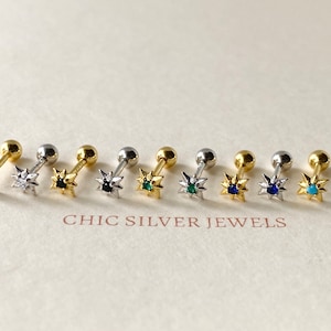 May include: Nine gold star-shaped earrings with different coloured gemstones in the centre. The earrings are on a white surface with the text "CHIC SILVER JEWELS".