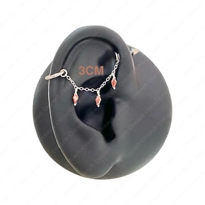 May include: Silver ear piercing jewelry with a chain and three dangling red gems. The chain measures 3 cm in length. The jewelry is designed to be worn on the ear.