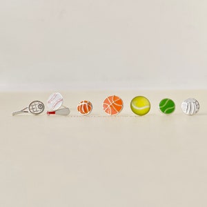 May include: A set of six silver earrings featuring sports-themed charms. The charms include a tennis racket, baseball and bat, baseball, basketball, tennis ball, and volleyball.