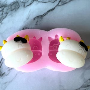 Puede incluir: Molde de silicona rosa con dos cavidades en forma de caras de vaca. El molde está lleno de caras de vaca de fondant blanco con ojos negros y cuernos amarillos.