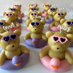 May include: A group of small, brown teddy bear candles with purple and pink heart-shaped sunglasses. Each bear is sitting in a purple or pink inflatable ring with a duck design.