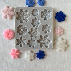 May include: A silicone mold with 12 snowflake designs. The mold is gray and the snowflakes are outlined in white. There are several finished snowflakes in various colors, including pink, blue, and white, surrounding the mold.