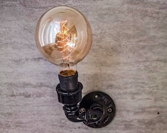 Könnte beinhalten: Ein schwarzer Metallrohranschluss mit einer durchsichtigen Glühbirne. Die Glühbirne hat ein sichtbares Filament, wenn sie leuchtet.