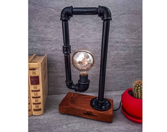 May include: A black industrial pipe table lamp with a wood base and a clear glass light bulb. The lamp is on a grey surface with a book and a potted plant.