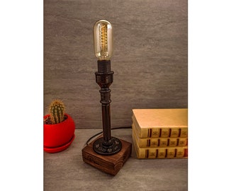 May include: A black metal table lamp with a vintage Edison bulb. The lamp is on a wooden base and has a pipe-like design. The lamp is sitting on a table with a stack of books.