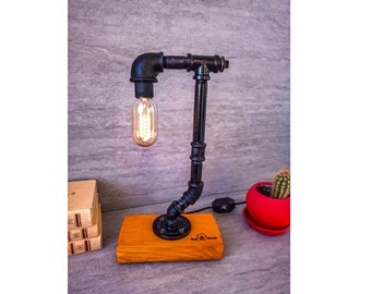 Peut inclure: Lampe de table de style industriel noir avec une ampoule Edison et une base en bois. La lampe est faite de tuyaux en métal noir et a un design simple.