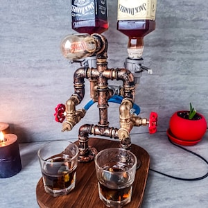 Puede incluir: Un dispensador de licor de estilo steampunk con dos botellas de whisky Jack Daniel's Tennessee, una normal y una de miel, montadas en un marco de tuber&iacute;a de cobre. El dispensador tiene dos v&aacute;lvulas rojas y dos vasos de chupito de vidrio transparente llenos de l&iacute;quido &aacute;mbar sobre una base de madera.