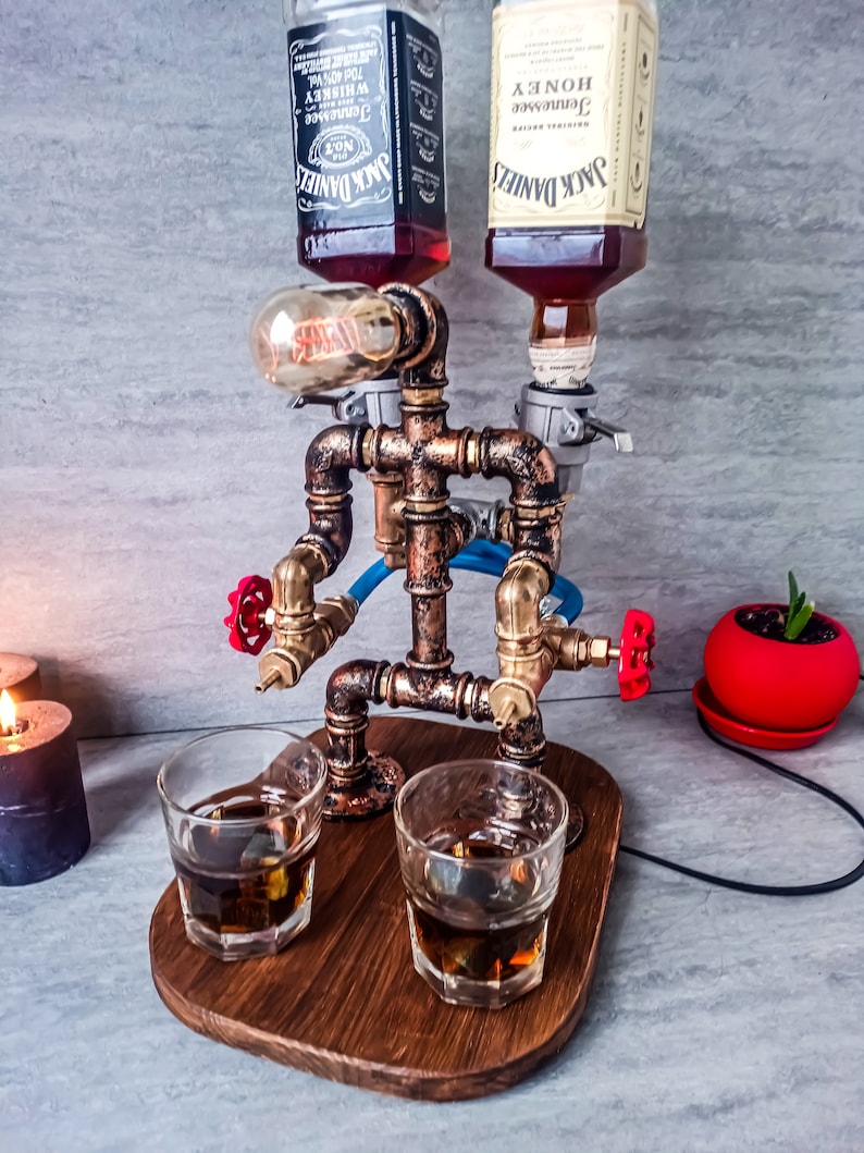 Puede incluir: Un dispensador de licor de estilo steampunk con dos botellas de whisky Jack Daniel's Tennessee, una normal y una de miel, montadas en un marco de tuber&iacute;a de cobre. El dispensador tiene dos v&aacute;lvulas rojas y dos vasos de chupito de vidrio transparente llenos de l&iacute;quido &aacute;mbar sobre una base de madera.