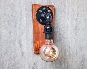 Könnte beinhalten: Eine schwarze Metall-Rohrleuchte mit einer Holzrückwand, montiert an einer grauen Wand. Die Leuchte hat eine klare Glühbirne mit einem Filament.
