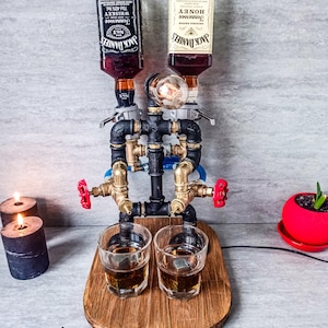 Puede incluir: Un dispensador de licor de metal negro y lat&oacute;n con dos botellas de vidrio de Jack Daniel's Tennessee Whiskey y Jack Daniel's Tennessee Honey. El dispensador tiene dos v&aacute;lvulas rojas y dos vasos de chupito de vidrio transparente con cubitos de hielo sobre una base de madera.