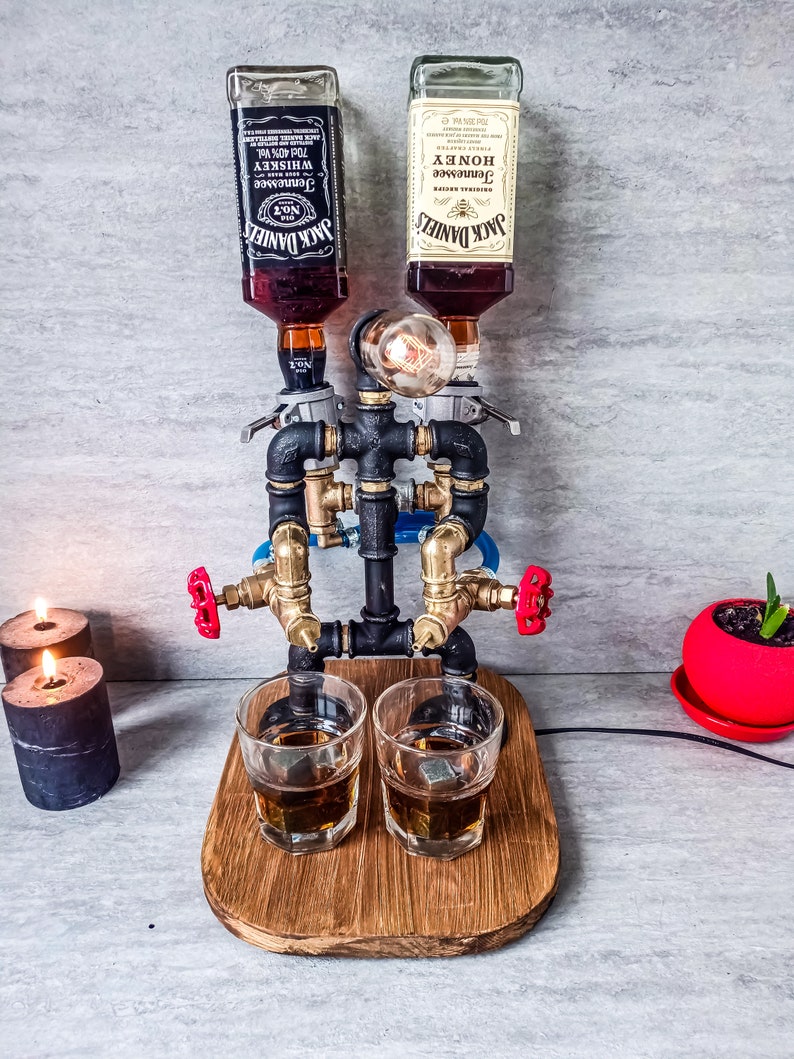 Puede incluir: Un dispensador de licor de metal negro y lat&oacute;n con dos botellas de vidrio de Jack Daniel's Tennessee Whiskey y Jack Daniel's Tennessee Honey. El dispensador tiene dos v&aacute;lvulas rojas y dos vasos de chupito de vidrio transparente con cubitos de hielo sobre una base de madera.