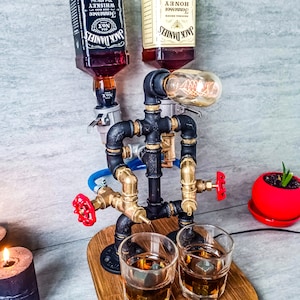 Puede incluir: Un dispensador de metal negro con tuber&iacute;as y v&aacute;lvulas, con dos botellas de Jack Daniel's Tennessee Whiskey y Honey, una bombilla y dos vasos con cubitos de hielo sobre una base de madera.