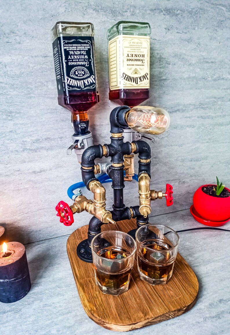 Puede incluir: Un dispensador de metal negro con tuber&iacute;as y v&aacute;lvulas, con dos botellas de Jack Daniel's Tennessee Whiskey y Honey, una bombilla y dos vasos con cubitos de hielo sobre una base de madera.