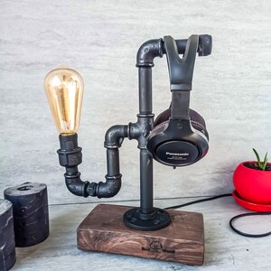 Puede incluir: Soporte de tuber&iacute;a de metal negro con una base de madera, que sostiene unos auriculares negros y una bombilla. La base est&aacute; grabada con "Red Wood".