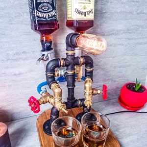 Puede incluir: Un dispensador de licor DIY hecho de tuber&iacute;as negras y de lat&oacute;n con dos botellas de vidrio de Jack Daniel's Tennessee Whiskey y Jack Daniel's Tennessee Honey Whiskey. El dispensador tiene dos v&aacute;lvulas rojas y una bombilla. Dos vasos con cubitos de hielo est&aacute;n sobre una base de madera.
