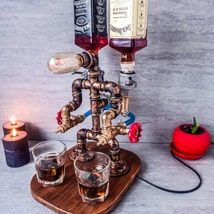 Puede incluir: Un dispensador de licor de estilo steampunk con dos botellas de Jack Daniel's, una normal y una de miel, montadas en un marco de tuber&iacute;a de cobre. El dispensador tiene dos boquillas y una bombilla. Dos vasos de licor est&aacute;n sobre una base de madera.
