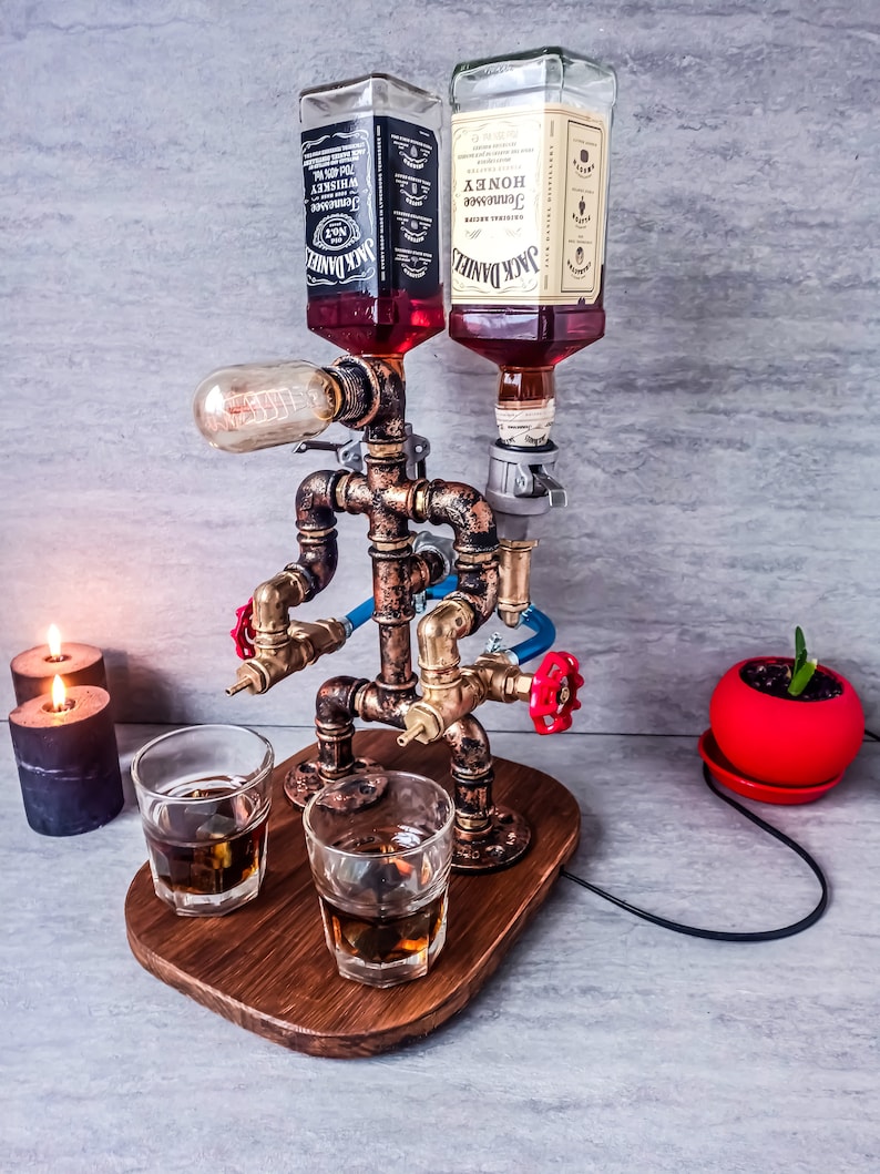 Puede incluir: Un dispensador de licor de estilo steampunk con dos botellas de Jack Daniel's, una normal y una de miel, montadas en un marco de tuber&iacute;a de cobre. El dispensador tiene dos boquillas y una bombilla. Dos vasos de licor est&aacute;n sobre una base de madera.
