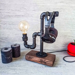 Puede incluir: Un soporte para auriculares de metal negro con una bombilla y una base de madera. El soporte est&aacute; hecho de tuber&iacute;as y tiene un aspecto industrial r&uacute;stico. La base es de madera oscura y tiene las palabras "Real Wood" grabadas.