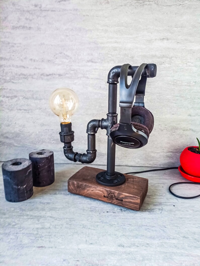 Puede incluir: Un soporte para auriculares de metal negro con una bombilla y una base de madera. El soporte est&aacute; hecho de tuber&iacute;as y tiene un aspecto industrial r&uacute;stico. La base es de madera oscura y tiene las palabras "Real Wood" grabadas.