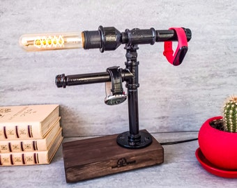 Peut inclure: Une lampe en métal noir avec une base en bois et une ampoule. La lampe a un support pour montre et un support pour téléphone. La lampe est sur une table avec une pile de livres et un pot rouge avec un cactus.