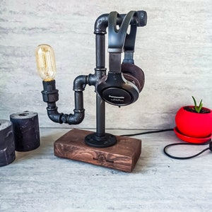 Puede incluir: Un soporte para auriculares de metal negro con una bombilla y una base de madera. El soporte est&aacute; hecho de tuber&iacute;as industriales y tiene un aspecto r&uacute;stico. Los auriculares son negros con la palabra "Panasonic" impresa en ellos.