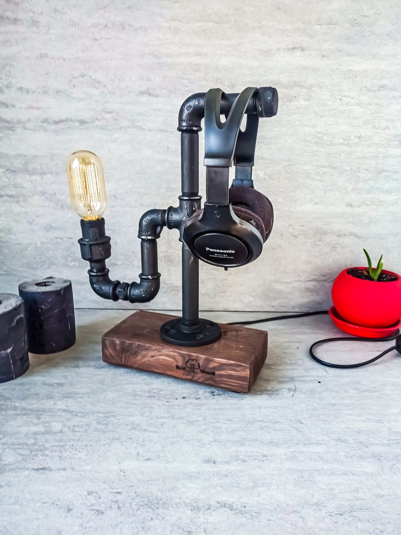 Puede incluir: Un soporte para auriculares de metal negro con una bombilla y una base de madera. El soporte est&aacute; hecho de tuber&iacute;as industriales y tiene un aspecto r&uacute;stico. Los auriculares son negros con la palabra "Panasonic" impresa en ellos.