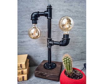Peut inclure: Une lampe de table en métal noir avec deux ampoules Edison. La lampe est faite de tuyaux industriels et a une base en bois. La lampe est sur une table avec une pile de livres et un cactus en pot.