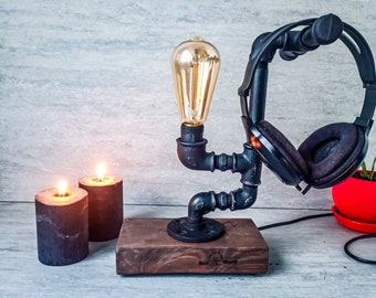 Puede incluir: Un soporte para auriculares de metal negro con una bombilla y una base de madera. El soporte está hecho de tuberías industriales y tiene un aspecto rústico. La base es de madera oscura y tiene las palabras "Real Wood" grabadas.