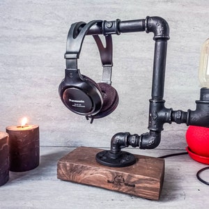 Op de afbeelding: Een zwarte koptelefoonstandaard gemaakt van industriële buizen met een houten basis. De standaard heeft een gloeilamp eraan bevestigd en houdt een paar zwarte koptelefoons vast. De standaard staat op een tafel met twee zwarte kaarsen.