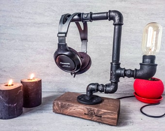 Puede incluir: Un soporte para auriculares negro hecho de tuberías industriales con una base de madera. El soporte tiene una bombilla conectada y sostiene un par de auriculares negros. El soporte está sobre una mesa con dos velas negras.