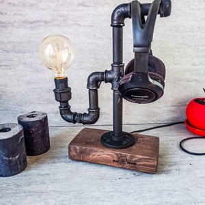 Puede incluir: Un soporte de tuber&iacute;a de metal negro con una base de madera, dise&ntilde;ado para sostener auriculares y una bombilla. El soporte cuenta con una bombilla negra y unos auriculares negros con la palabra "Panasonic" impresa en el lateral. La base de madera tiene las palabras "Red Wood" grabadas.