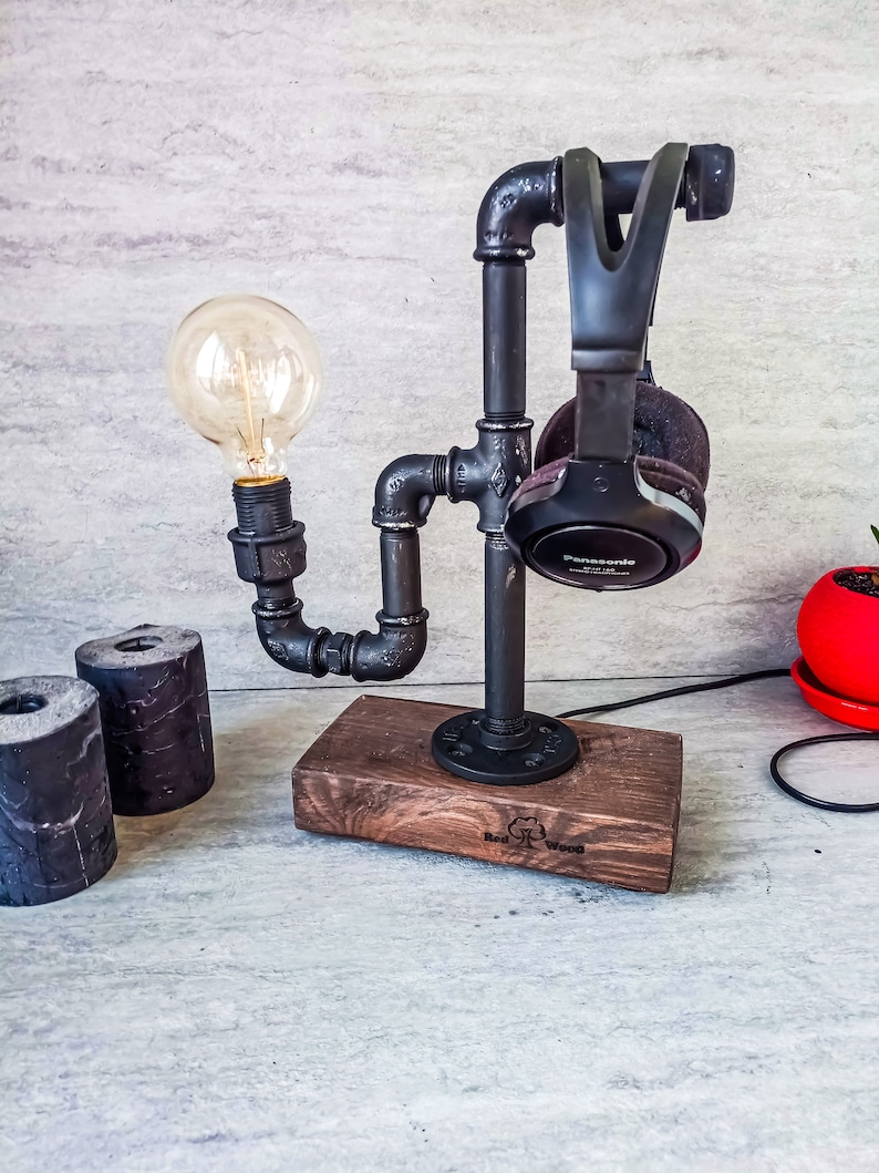 Puede incluir: Un soporte de tuber&iacute;a de metal negro con una base de madera, dise&ntilde;ado para sostener auriculares y una bombilla. El soporte cuenta con una bombilla negra y unos auriculares negros con la palabra "Panasonic" impresa en el lateral. La base de madera tiene las palabras "Red Wood" grabadas.