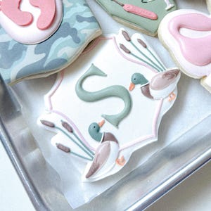 May include: Decorated sugar cookies in a metal baking pan. One cookie features a white background with a pink and green border, a green letter "S", and two ducks. Other cookies have a camouflage pattern, baby footprints, and a pink swan.