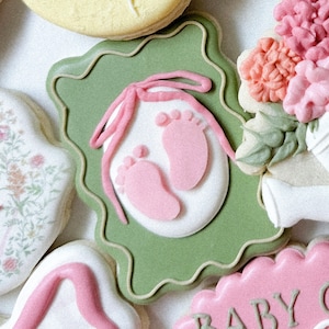 Op de afbeelding: Een groene en witte koek versierd met roze babyvoetjes en een roze lint. De koek is in de vorm van een fotolijst.