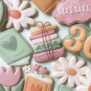 May include: An assortment of decorated cookies in pastel colors. The cookies include shapes of books, flowers, and the number 30. Some cookies have the word "Autumn" on them. The cookies are decorated with pink, green, and orange icing.