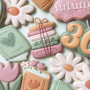 以下が含まれることがあります： パステルカラーでデコレーションされたクッキーの詰め合わせ。クッキーには、デイジー、本、紐で縛られた本の山、数字の30、そして「Autumn」と書かれた看板が含まれています。クッキーはピンク、緑、オレンジ色のアイシングで飾られています。