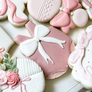May include: A set of pink and white decorated sugar cookies with various designs, including a baby onesie, a bow, and a flower.