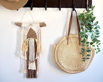 Puede incluir: Un sombrero marrón, un bolso tejido y un tapiz de macramé con borlas marrones y blancas cuelgan de un perchero de metal negro con cinco ganchos.