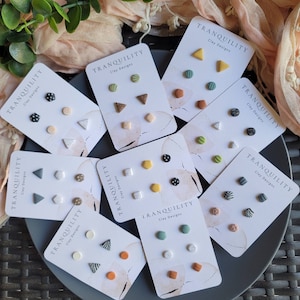 Op de afbeelding: Assortiment van acht sets kleioorbellen, elk set met drie verschillende ontwerpen. De oorbellen worden gepresenteerd op een grijs bord met een witte achtergrond. De sets bevatten geometrische vormen, stippen, strepen en andere patronen. Elk set is gelabeld met "TRANQUILITY Clay Designs".