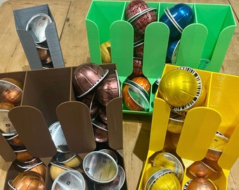 Op de afbeelding: Verschillende 3D-geprinte koffiepadhouders in grijs, bruin, groen en geel. Elke houder heeft meerdere compartimenten en is gevuld met metallic koffiepads. Ontworpen om koffiecapsules in op te bergen.