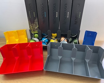 Op de afbeelding: Een verzameling Nespresso koffiecapsules en -organizers. De afbeelding toont verschillende zwarte Nespresso dozen met verschillende koffievariëteiten. Er zijn ook diverse 3D-geprinte organizers in rood, geel, grijs en blauw, ontworpen om de capsules te bewaren.