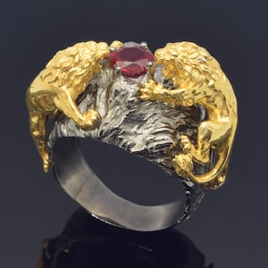 May include: A gold ring with two lion figures on either side of a large red gemstone. The ring is made of silver and gold.