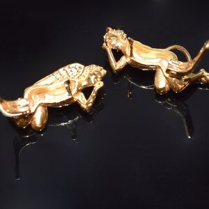 May include: Two gold-toned angel figurines with wings, lying on their sides, with one angel's head resting on its hand. The figurines are on a black surface.