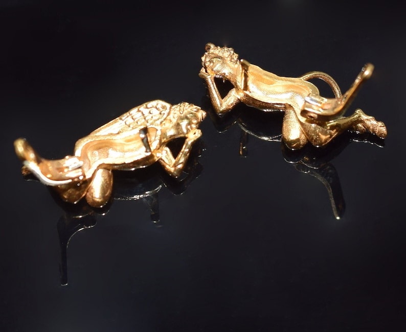 May include: Two gold-toned angel figurines with wings, lying on their sides, with one angel's head resting on its hand. The figurines are on a black surface.