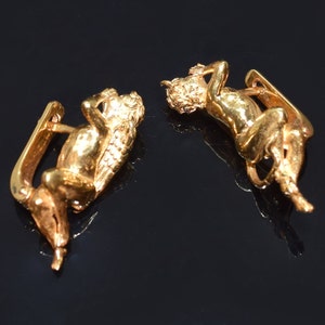 May include: A pair of gold-tone earrings featuring a cherub design. Each earring has a cherub figure with wings, lying down and holding its head in its hand.